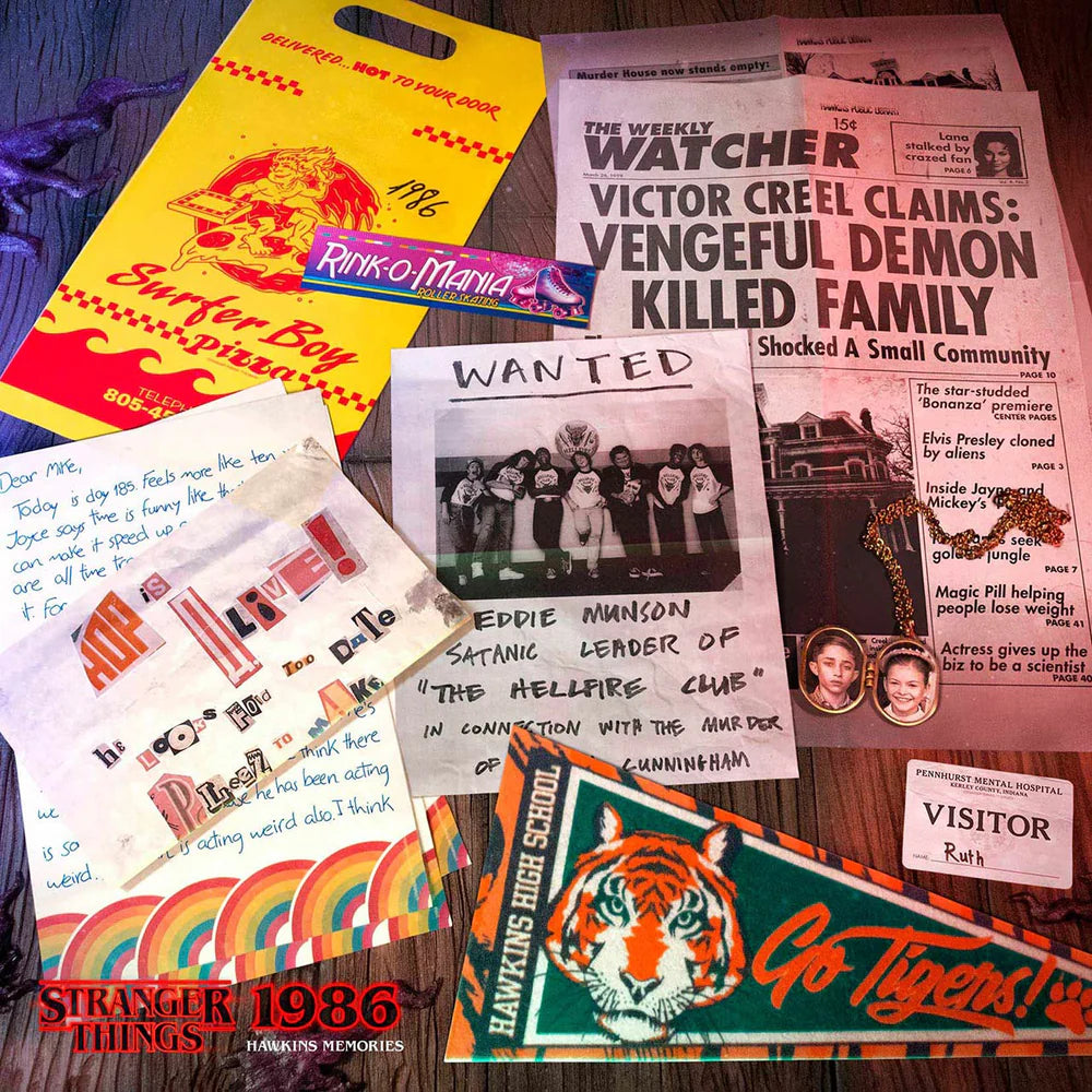 A flat lay of various Strange Things / Marvel Comics memorabilia items, including a yellow and red 'Stranger Boy' bag, a 'The Watcher' newspaper clipping, a handwritten letter, a 'Wanted' poster, a 'Visitor' card, and a 'Go Tigers!' sticker, all arranged on a dark wooden surface.